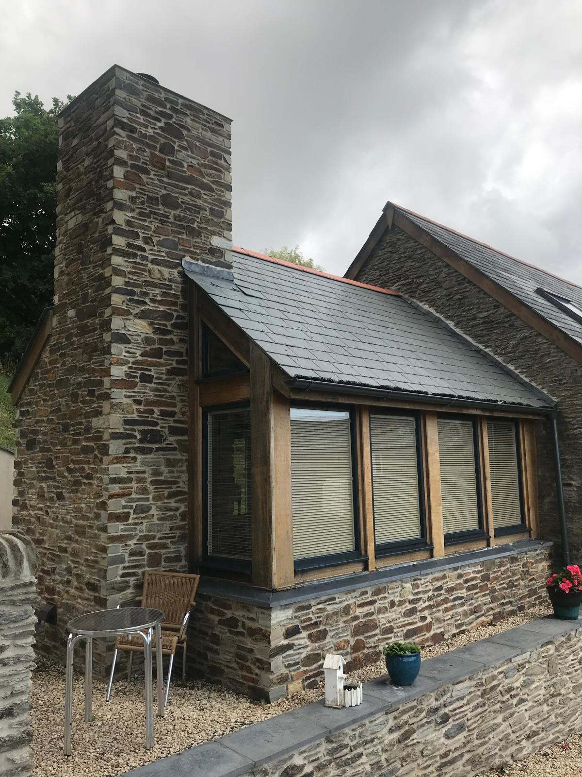 A stone house with a large chimney, slate roof, and corner windows with wooden frames showcases the charm of barn conversions. In front, there’s a small patio with a table, two chairs, and potted plants. The sky is overcast.