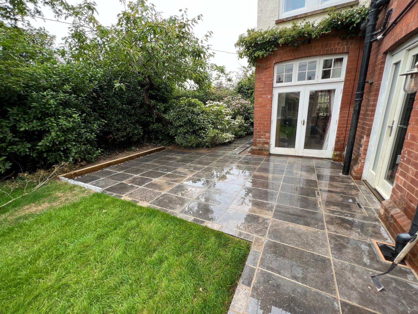 A patio with dark, wet tiles showcases quality stone masonry beside green grass and lush bushes, all adjacent to a red brick house with white-framed glass doors and windows.