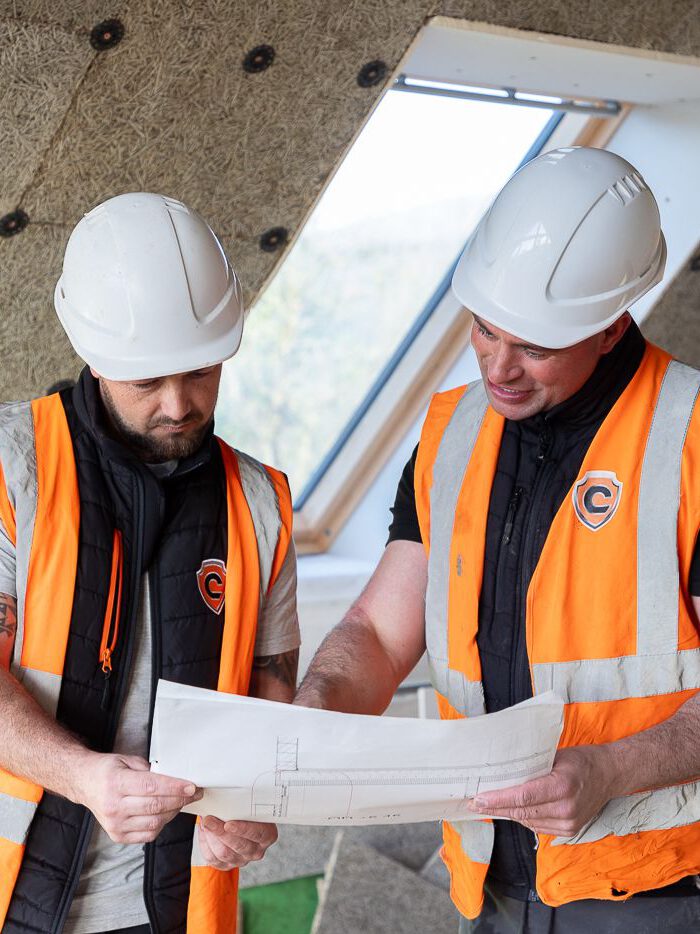 Two builders in orange safety vests and white helmets review building plans inside a partially completed structure, with insulation materials in the background—an essential step for quality barn conversions or Passivhaus buildings.